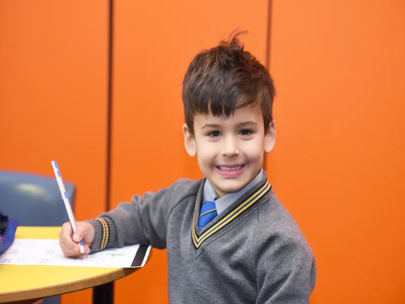 A young Coogee Prep boy smiling at his desk with a pencil in hand